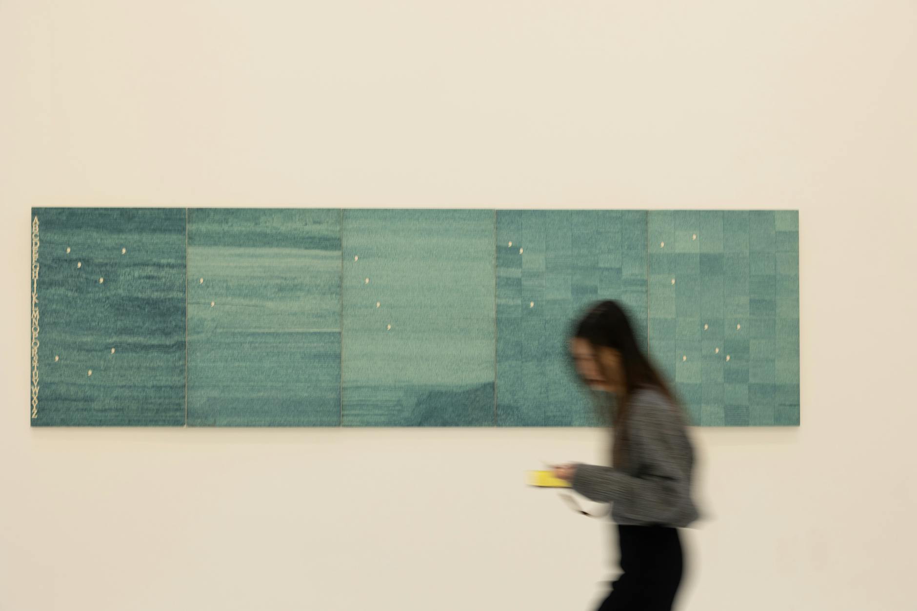 현대적인 분위기의 갤러리 내부에서 추상화 작품 앞을 지나가는 관람객의 모습입니다.