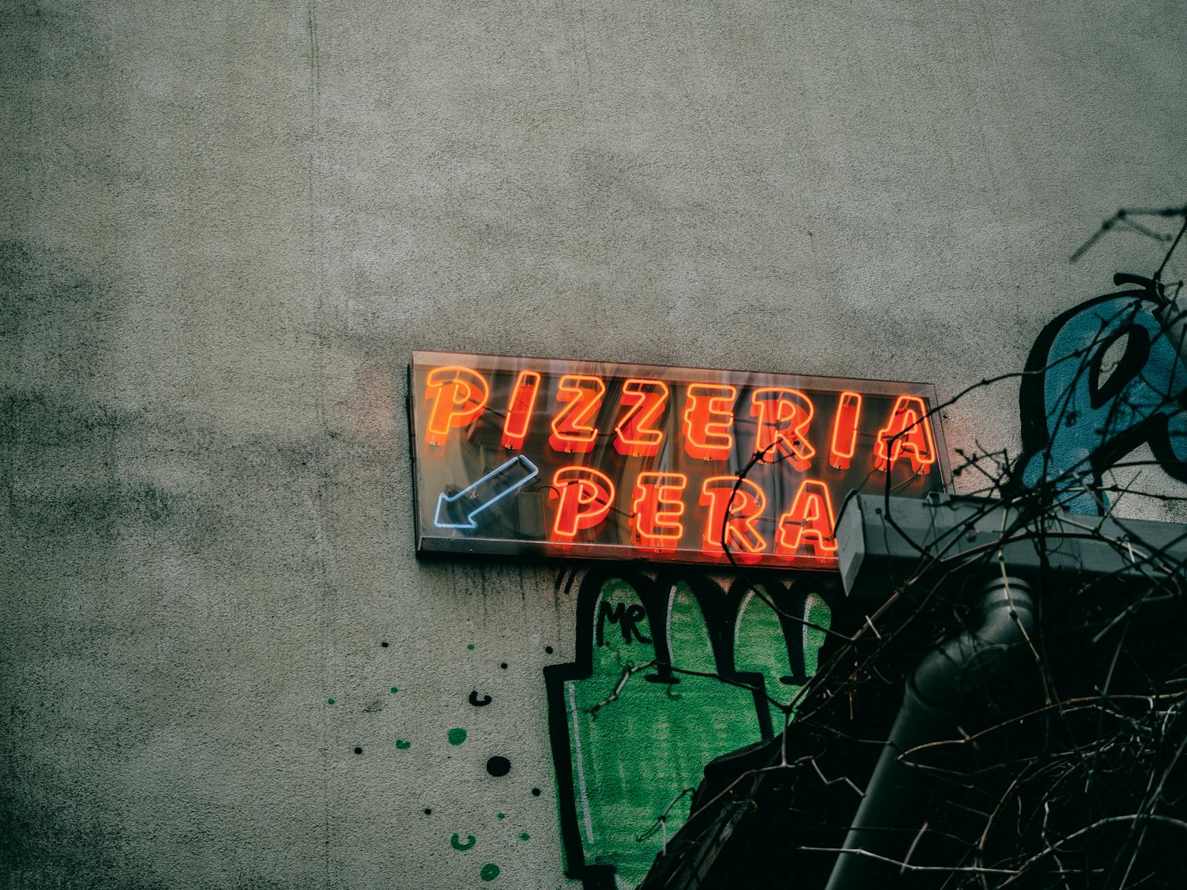 낡은 콘크리트 벽에 걸린 주황색 네온사인 'PIZZERIA PERA'가 희미하게 빛나고 있습니다. 이 이미지는 [투어] 시간이 멈춘 골목을 걷다: 15년 거주자 Baran이 안내하는 추쿠르주마 골동품 & 페라 비밀 통로 투어와 관련된 이스탄불의 오래된 거리 분위기를 보여주며, 파이프와 덩굴로 일부 가려져 있습니다.